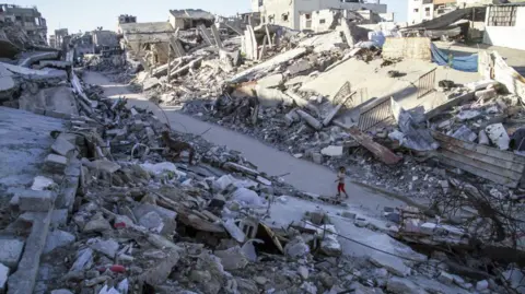 Getty Images A wide shot of Jabalia, where a child stands on a road where either side of him is grey rubble.