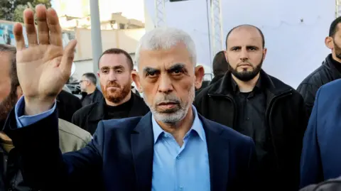 Yahya Sinwar waves as he looks towards the camera wearing a suit and blue shirt with other men in the background, while addressing Palestinians in Gaza in April